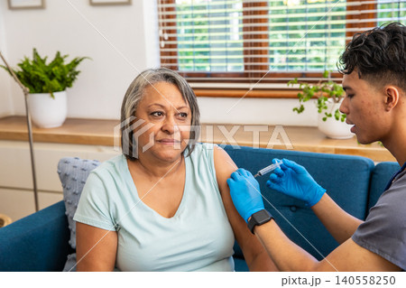 Senior female getting vaccine injection using syringe from asian clinician on sofa 140558250