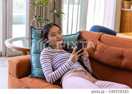 Asian woman reclining on rust-colored sofa near sliding glass door and plant holding smartphone 140558383