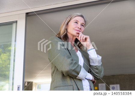 Senior woman wearing green blazer standing on patio, wall beside glass door Senior woman wearing green blazer standing on patio, wall beside glass door 140558384