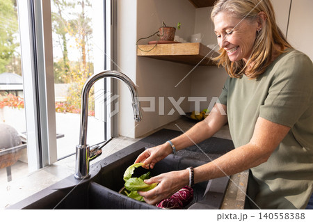 Senior woman washing lettuce and radicchio under chrome faucet at sink by window 140558388