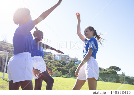Diverse teenage teammates celebrating on soccer field by goal wearing blue jerseys and shorts 140558410