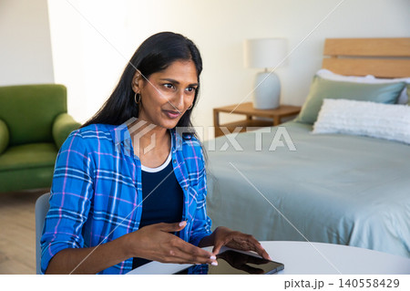 Indian woman sitting at round white table near bed, using smartphone in bedroom, copy space Indian woman sitting at round white table near bed, using smartphone in bedroom, copy space 140558429