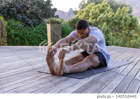 Man stretching on exercise mat on deck with planter wearing white t-shirt and black shorts Man stretching on exercise mat on deck with planter wearing white t-shirt and black shorts 140558440