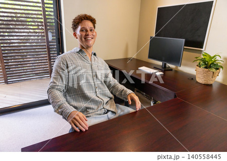 African american man sitting at office desk smiling at camera with computer monitor and plant African american man sitting at office desk smiling at camera with computer monitor and plant 140558445