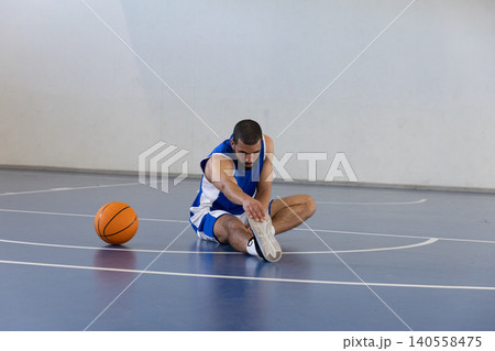 Male basketball player in blue uniform sitting stretching leg on basketball court near ball 140558475