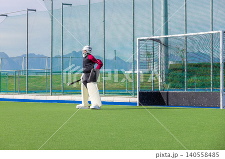 Male field hockey goalkeeper in protective gear guarding goal on synthetic turf holding stick Male field hockey goalkeeper in protective gear guarding goal on synthetic turf holding stick 140558485