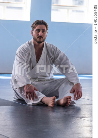 Man sitting cross-legged on padded floor in dojo in white martial arts gi under bright windows 140558488