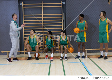 Diverse male basketball team listening to coach shouting in gym with basketballs and bench Diverse male basketball team listening to coach shouting in gym with basketballs and bench 140558492