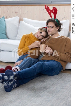 Couple sitting on grey rug in living room holding beige mugs and wearing red antler headbands Couple sitting on grey rug in living room holding beige mugs and wearing red antler headbands 140558517