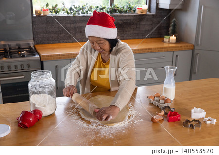 Senior Asian woman wearing Santa hat rolling cookie dough on kitchen countertop with flour jar Senior Asian woman wearing Santa hat rolling cookie dough on kitchen countertop with flour jar 140558520