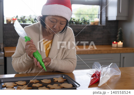 Senior Asian woman wearing Santa hat decorating cookies using green icing bag at kitchen counter Senior Asian woman wearing Santa hat decorating cookies using green icing bag at kitchen counter 140558524