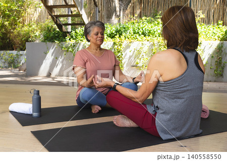 Senior and middle-aged female friends meditating on deck yoga mats by gray bottle and white towel Senior and middle-aged female friends meditating on deck yoga mats by gray bottle and white towel 140558550