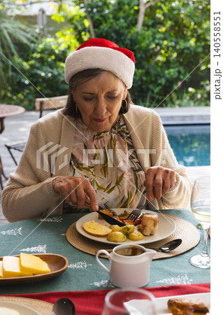 Senior woman wearing Santa hat cutting holiday meal at patio beside pool with plate of food 140558551