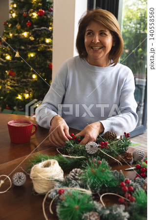 Senior woman sitting at table in living room making holiday wreath with pinecones and red mug Senior woman sitting at table in living room making holiday wreath with pinecones and red mug 140558560