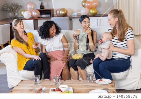 Diverse female friends feeding baby strawberry on sofa with chips, balloons in living room Diverse female friends feeding baby strawberry on sofa with chips, balloons in living room 140558578