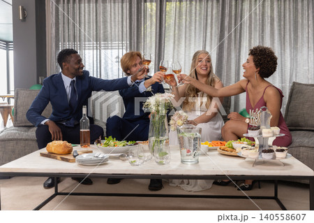 Diverse friends sitting on couch raising rosé wine glasses in living room, coffee table Diverse friends sitting on couch raising rosé wine glasses in living room, coffee table 140558607