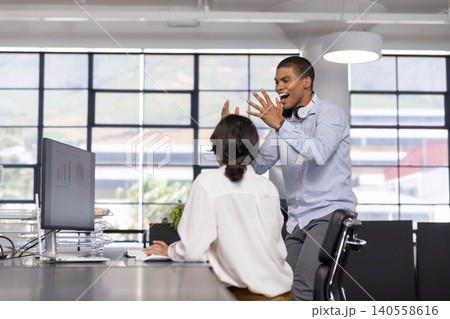 Diverse coworkers discussing report at office desk with monitor, plant, headphones, copy space Diverse coworkers discussing report at office desk with monitor, plant, headphones, copy space 140558616