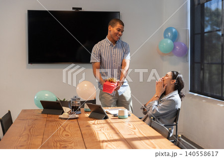 Coworkers presenting red gift box on conference table with laptops and balloons 140558617