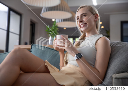 Woman holding mug on gray sofa in living room wearing smartwatch under pendant lamps 140558660