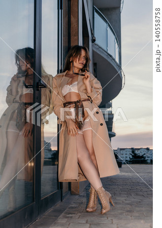 Trench coat lingerie woman leaning against glass door on terrace during sunset showcasing seductive fashion and urban elegance Trench coat lingerie woman leaning against glass door on terrace during sunset showcasing seductive fashion and urban elegance 140558758