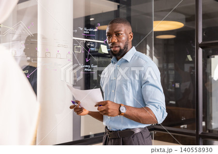 African american man drawing on glass whiteboard while holding papers and marker in office African american man drawing on glass whiteboard while holding papers and marker in office 140558930