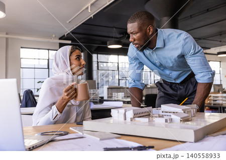 Diverse coworkers discussing architectural model beside laptop in open-plan office with coffee Diverse coworkers discussing architectural model beside laptop in open-plan office with coffee 140558933