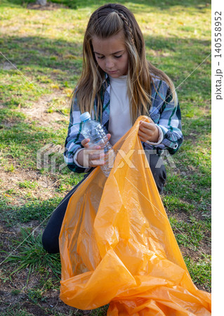 Girl kneeling in grassy field with clear plastic water bottle opening orange trash bag Girl kneeling in grassy field with clear plastic water bottle opening orange trash bag 140558952