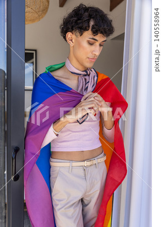 Man in twenties standing in home doorway with pride flag on shoulders and scarf 140558964
