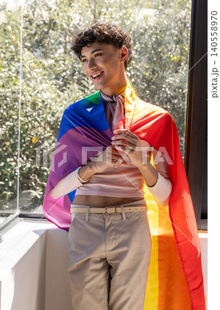 Man leaning on windowsill wearing pink sweater and rainbow pride flag by window 140558970
