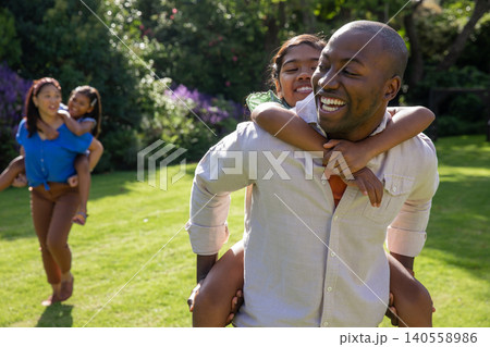 Diverse family walking across backyard lawn carrying daughters on backs in sunny garden Diverse family walking across backyard lawn carrying daughters on backs in sunny garden 140558986