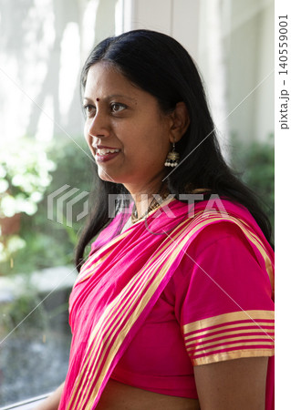 Indian woman wearing pink sari jhumka earrings and standing by glass window gazing at garden view Indian woman wearing pink sari jhumka earrings and standing by glass window gazing at garden view 140559001
