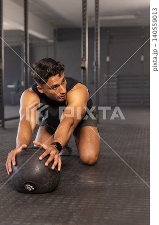 Man in sportswear kneeling on rubber mat holding 15-pound medicine ball and extending arms in gym Man in sportswear kneeling on rubber mat holding 15-pound medicine ball and extending arms in gym 140559013