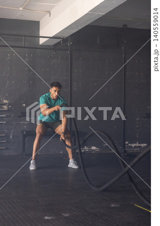 Male athlete performing battle rope exercise while sitting on bench in minimalist gym 140559014