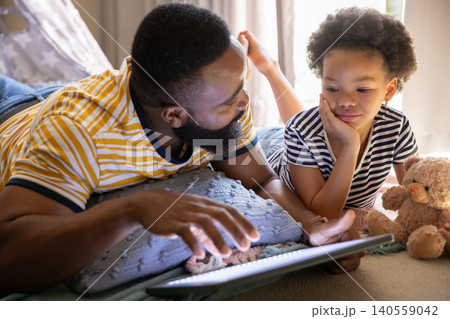 African American father and daughter lying on bed at home holding tablet alongside plush teddy bear 140559042