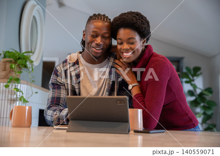 African american couple leaning together at kitchen table, browsing tablet with coffee mugs African american couple leaning together at kitchen table, browsing tablet with coffee mugs 140559071