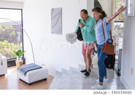 Diverse senior couple entering home foyer carrying travel bags, rolling suitcase, copy space 140559080