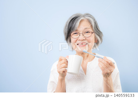 青色背景で歯ブラシを持って歯磨きによる口腔ケアを行うシニア女性 カメラ目線笑顔8020運動のイメージ 青色背景で歯ブラシを持って歯磨きによる口腔ケアを行うシニア女性 カメラ目線笑顔8020運動のイメージ 140559133