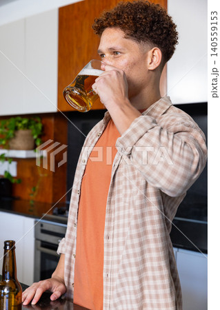 Man standing at kitchen stone countertop sipping beer from glass mug near bottle by plant 140559153