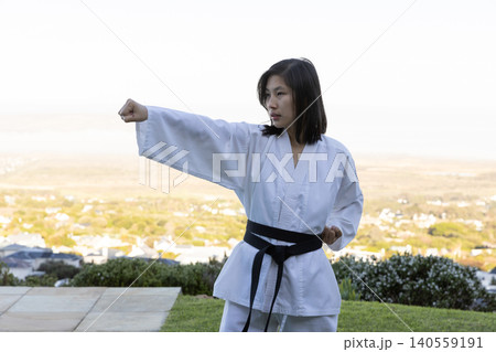 Asian female holding punching stance in gi and black belt on grass beside tiled patio Asian female holding punching stance in gi and black belt on grass beside tiled patio 140559191