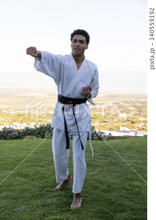 African american man punching barefoot in white gi and belt on grassy slope above suburbs African american man punching barefoot in white gi and belt on grassy slope above suburbs 140559192