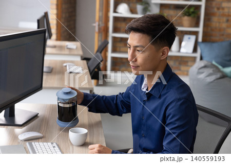 Asian man brewing coffee with french press at office desk with monitor, mug Asian man brewing coffee with french press at office desk with monitor, mug 140559193