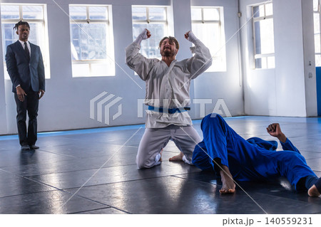 Diverse martial arts competitors and referee celebrating victory, observing match on padded mats Diverse martial arts competitors and referee celebrating victory, observing match on padded mats 140559231