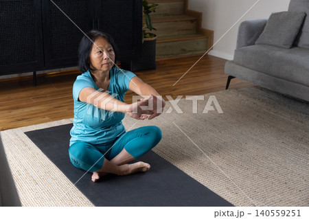 Middle-aged Asian woman stretching arms forward sitting on yoga mat at home in turquoise activewear Middle-aged Asian woman stretching arms forward sitting on yoga mat at home in turquoise activewear 140559251