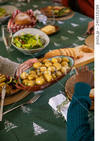 Diverse friends in sweaters passing brown ceramic dish of potatoes at home dining table Diverse friends in sweaters passing brown ceramic dish of potatoes at home dining table 140559298