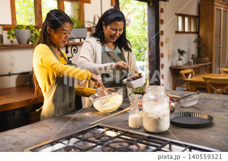 Diverse female friends wearing aprons whisking batter and adding cherries in home kitchen with bowl Diverse female friends wearing aprons whisking batter and adding cherries in home kitchen with bowl 140559321