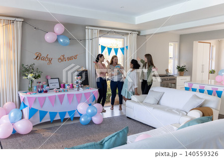Diverse female friends standing and chatting in living room under baby shower banner, balloons Diverse female friends standing and chatting in living room under baby shower banner, balloons 140559326