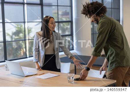 Diverse coworkers in business attire shaking hands at meeting table with laptop, tablet Diverse coworkers in business attire shaking hands at meeting table with laptop, tablet 140559347