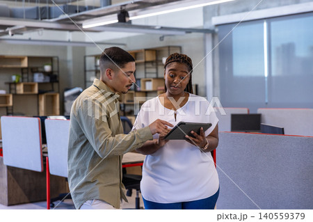 Diverse coworkers discussing details on tablet at desks in office with gray cubicle partitions Diverse coworkers discussing details on tablet at desks in office with gray cubicle partitions 140559379
