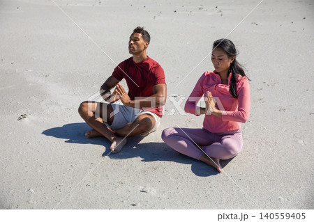 Diverse couple in sportswear sitting cross-legged on white beach meditating under sunlight Diverse couple in sportswear sitting cross-legged on white beach meditating under sunlight 140559405