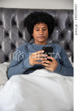 African american man reclining in bed at home using smartphone, tufted leather headboard African american man reclining in bed at home using smartphone, tufted leather headboard 140559407